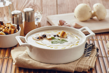 Mushroom cream soup on a table, with bread, garlic, and raw mushrooms on a white chopping board. Salt and Pepper on the table..
