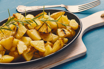 roasted potatoes iwith onion and rosemary in a cast iron pan over a white chopping board on a blue table