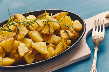roasted potatoes iwith onion and rosemary in a cast iron pan over a white chopping board on a blue table