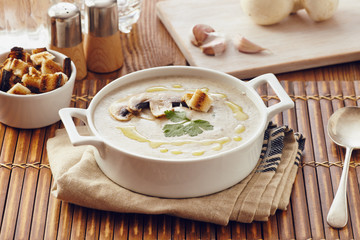 Mushroom cream soup on a table, with bread, garlic, and raw mushrooms on a white chopping board. Salt and Pepper on the table..