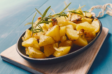 roasted potatoes iwith onion and rosemary in a cast iron pan over a white chopping board on a blue table