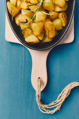 roasted potatoes iwith onion and rosemary in a cast iron pan over a white chopping board on a blue table