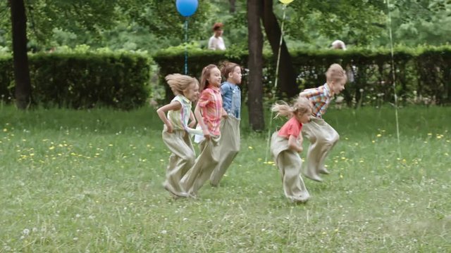 Slow motion shot of five boys and girls jumping in sacks through the green lawn in the park while having birthday party with parents cheering them