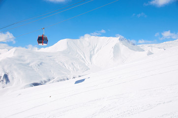 Fototapeta na wymiar Cable lift for skiers or snowboarders in brigth day on ski resort with blue sky and white snow mountain picks background. Copy space for a text. Red car or cabinet for athletes.