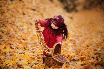 Toddler girl in Ukrainian or Russian folk scarf on head with floral print. Cute baby takes out many bagels from picnic basket in beautiful autumn forest thickly covered with orange fallen leaves.