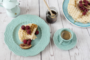Soft Belgian heart shaped waffles with raspberries and figs, covered with honey on turquoise blue plate. A coffee and coffee maker. Rustic wooden background, top view. Holiday morning at home