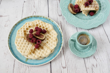 Soft Belgian heart shaped waffles with raspberries and figs, covered with honey on turquoise blue plate and coffee. Rustic wooden background, top view. Holiday morning at home