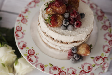 Naked cake with caramelized fruits - strawberries, blueberries, raspberries. Sponge cream cake in floral high plateau, tray. White roses, rustic background