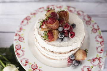 Naked cake with caramelized fruits - strawberries, blueberries, raspberries. Sponge cream cake in floral high plateau, tray. White roses, rustic background