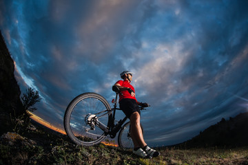 cyclist standing with mountain bike on trail at sunset
