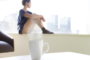 Beautiful woman looking at the window in the morning. Relaxing young woman awakening with a hot cup in the city
