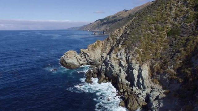 Aerial shot flying by the California Big Sur coast. Deep blue ocean and massive cliffs with waves crashing. Blue skies with sparse clouds.
