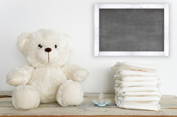 A teddy bear on a wooden table next to a pacifier and some diapers. A blackboard with empty copy space for Author's text.