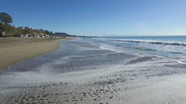 Low flying aerial shot over the waves and beach, taken in Santa Cruz California. Ocean beach houses visible with white foamy waves crashing below. Clear blue sky.
