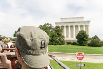 A tourist in a baseball cap, sits on an open deck bus in Washington DC on a cloudy summer's day...
