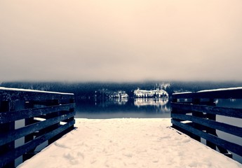 Snow covered wooden jetty on the lake; foggy winter landscape in black and white. Monochrome image filtered in retro, vintage style with soft focus and red filter. Lake Bohinj, Slovenia - 121616929