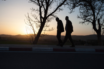 2 men in morocco are walking across the sunset, between trees, on a street.