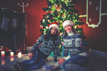 Young female friends in hats Santa Claus near a Christmas tree in the decorated room.