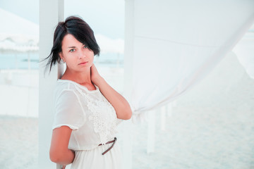 Woman, brunette with short hair in white dress on the beach of ocean, sea near sun shade, tent. Vacation.