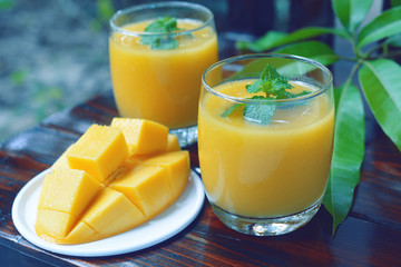 ripe yellow mango smoothie decorated by mint leaf on top with fruit and green leaf on wooden background.