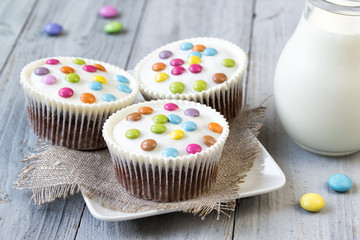 Obraz na płótnie Canvas Chocolate cupcakes with white icing and colored smarties on a plate and glass jar of milk, wooden background