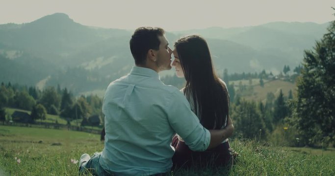 Lovers man and woman rubbing noses against each other on green plain. Men hugs her girlfriend and kisses on the forehead. 