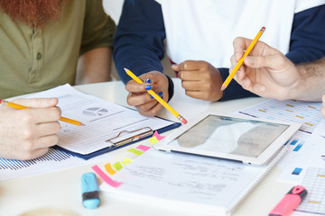 Team of creative young people holding pencils while discussing business strategy and plans of their collaborative project, using digital tablet, sharing vision and ideas, sitting at table in office