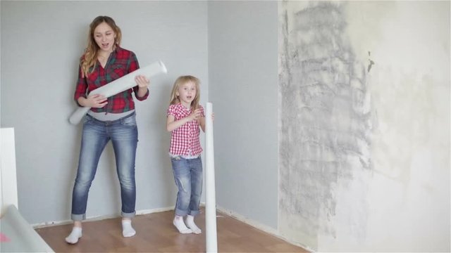 Cheerful girls are dancing and singing near the wall with rolls of wallpaper in their hands.