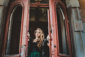 Beautiful girl in green vintage dress with coat walking near ancient house and door. Vintage farytale concept. Woman with long hair and make up