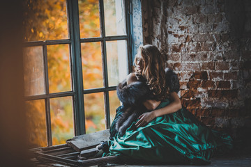 Fototapeta na wymiar young pretty women with long lush curly blonde hair and thoughtful face in green dress standing near stony wall and big window sunny day