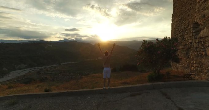 Giovane ragazzo sta mettendo le sue mani in aria di fronte a un tramonto oltre le montagne in una collina.