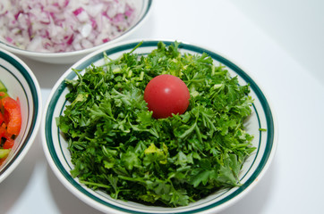 One bowl of cilantro-parsely surrounded by  chopped onions and peppers