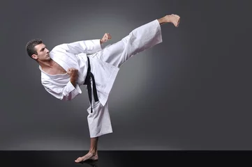 Fototapete Kampfkunst Hübscher junger schwarzer Gürtel männliches Karate, das Kick auf dem grauen Hintergrund macht