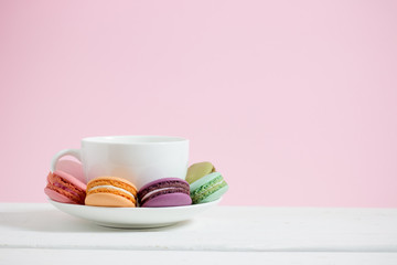 Colorful french macarons and white coffee cup on white table wooden pastel pink background.