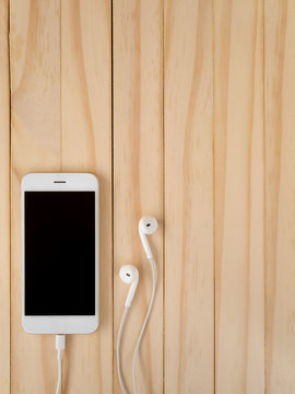 Front view image of new smartphone connecting with charging cable and earbuds on wooden background with copy space.
