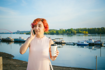 Fototapeta na wymiar girl in pale pink dress with red hair and backpack walking along river bank, talking on the phone and drinking coffee from a cardboard cup, against backdrop of boats moored on a warm summer day