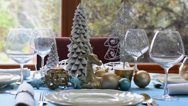 Stylish blue, gold and white Christmas table with flickering fairy lights centerpiece in front of garden window, panning across table.