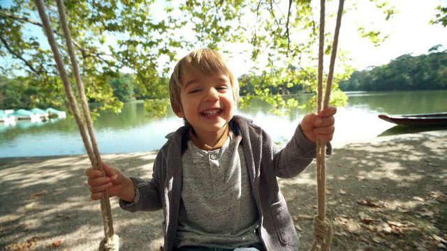 4k footage, 2 year old smiling boy swinging on swing on sunny day at lake
