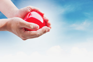 Hand holding red hart on blue sky and white clouds, with bright light