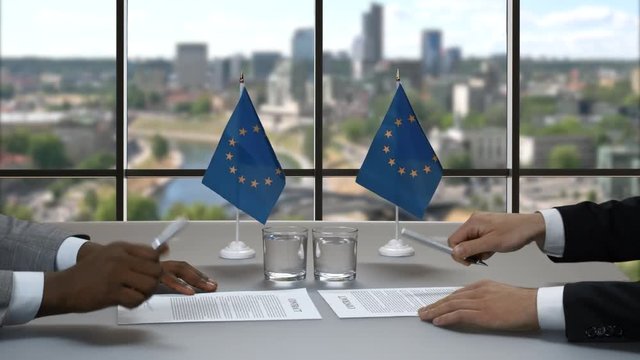 Businessmen sign papers at desk. Men shake hands beside flags. Policy of European Union. Time of big changes.