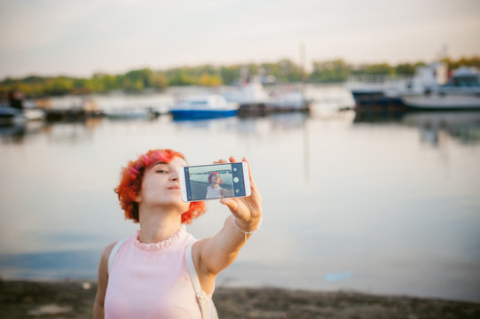 girl in pale pink dress with red hair and backpack walking along river bank, pictures of themselves on their mobile camera phone, against backdrop of boats moored on a warm summer day