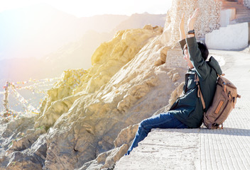 Extremely happy and joyful male traveler sitting and raising hands up in mountain area.