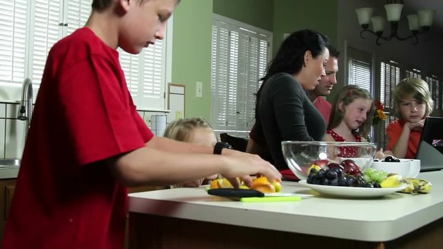 Big brother prepares healthy snack for little sister while rest of family looks at something on laptop.