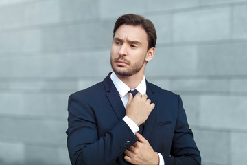 Elegant and successful. Confident young businessman correcting his necktie and looking away while standing outdoors 
