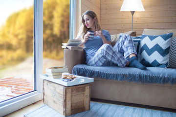 Naklejka premium Young beautiful blonde woman with cup of coffee and book sitting home by the window. Autumn fall trees view. Lazy day off concept