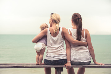 Loving mother embracing her children sitting on wooden bench at tropical beach during vacation. They together looking into the distance. Concept for togetherness and  bright future. - obrazy, fototapety, plakaty