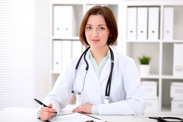 Fototapeta na wymiar Young brunette female doctor sitting at the table and working at hospital office. Health care, insurance and help concept. Physician ready to examine patient