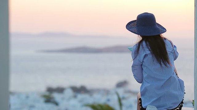 Young woman is looking at the sunset over a sea in famous village Mykonos with the old buildings in the background. Soft summer backlight.