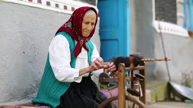 Elderly woman sits on bench and works with distaff with spinning wheel. Ukrainian old woman sits on bench near his house and spin yarn. Female with spinning wheel, handicraft