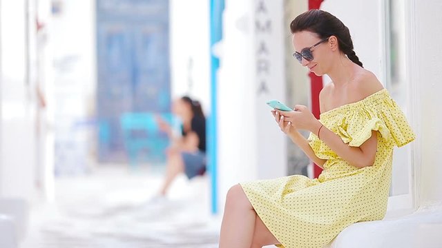 Beautiful woman with smart phone outdoors in old streets an Mykonos. Girl at street of typical greek traditional village with white walls and colorful doors on Mykonos Island, in Greece, Europe